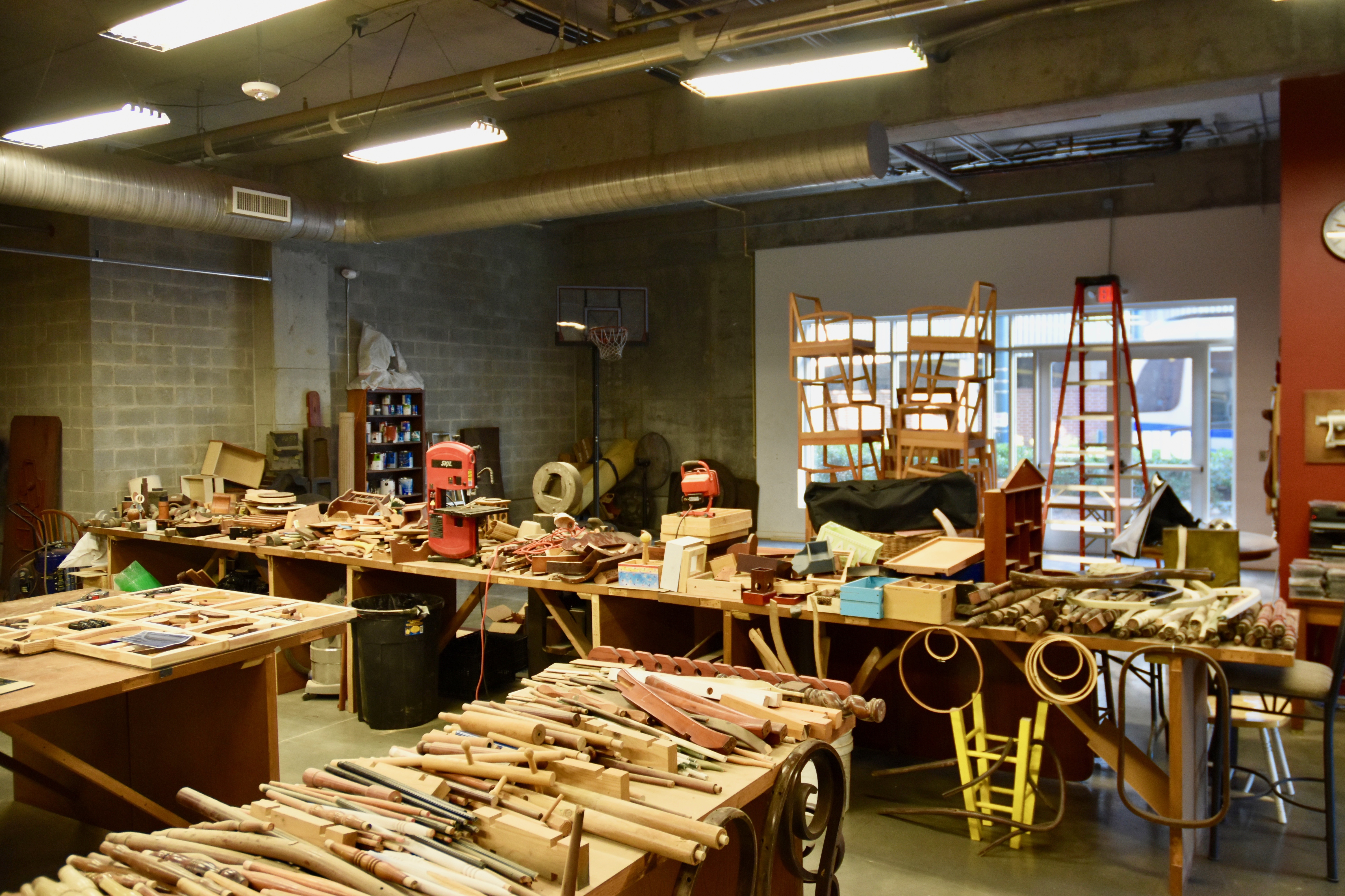 The inside of David Furman’s construction studio and exhibition space, a cluttered woodworking shop filled with various tools and materials, including reclaimed wooden pieces and furniture parts on multiple tables. Shelves in the background hold additional equipment, and a ladder leans against the wall with stacked chairs nearby.