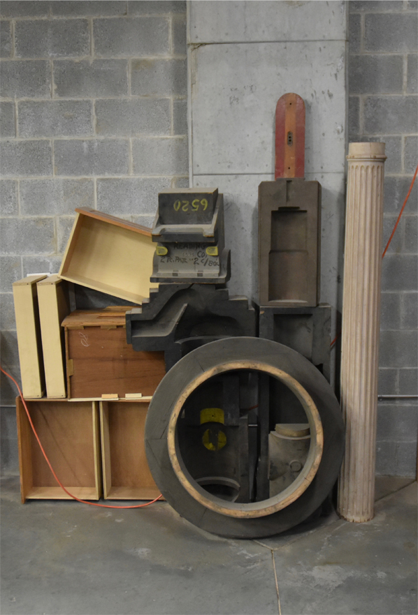 A collection of various reclaimed wooden materials stacked against a concrete wall, including a large circular piece, a column, and other shapes inside the studio of artis David Furman