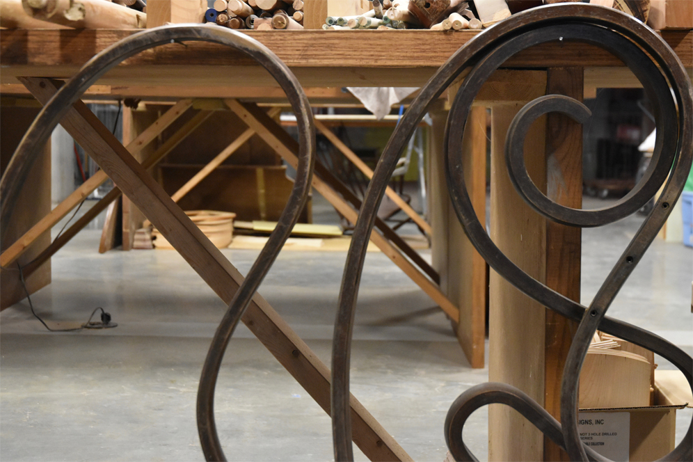 View inside David Furman workspace and studio depicting wooden table, featuring intricate curves and details, with reclaimed wooden items scattered on the table's surface.