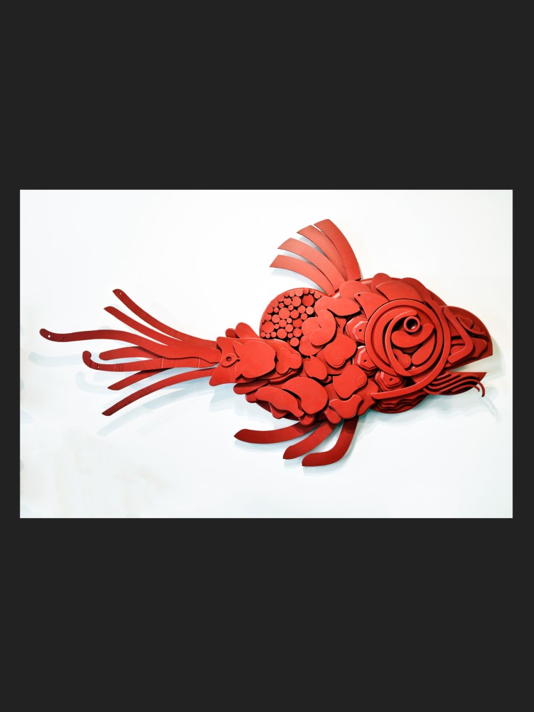A colorful abstract sculpture of a fish, made from reclaimed wood, featuring various geometric forms, painted red by David Furman