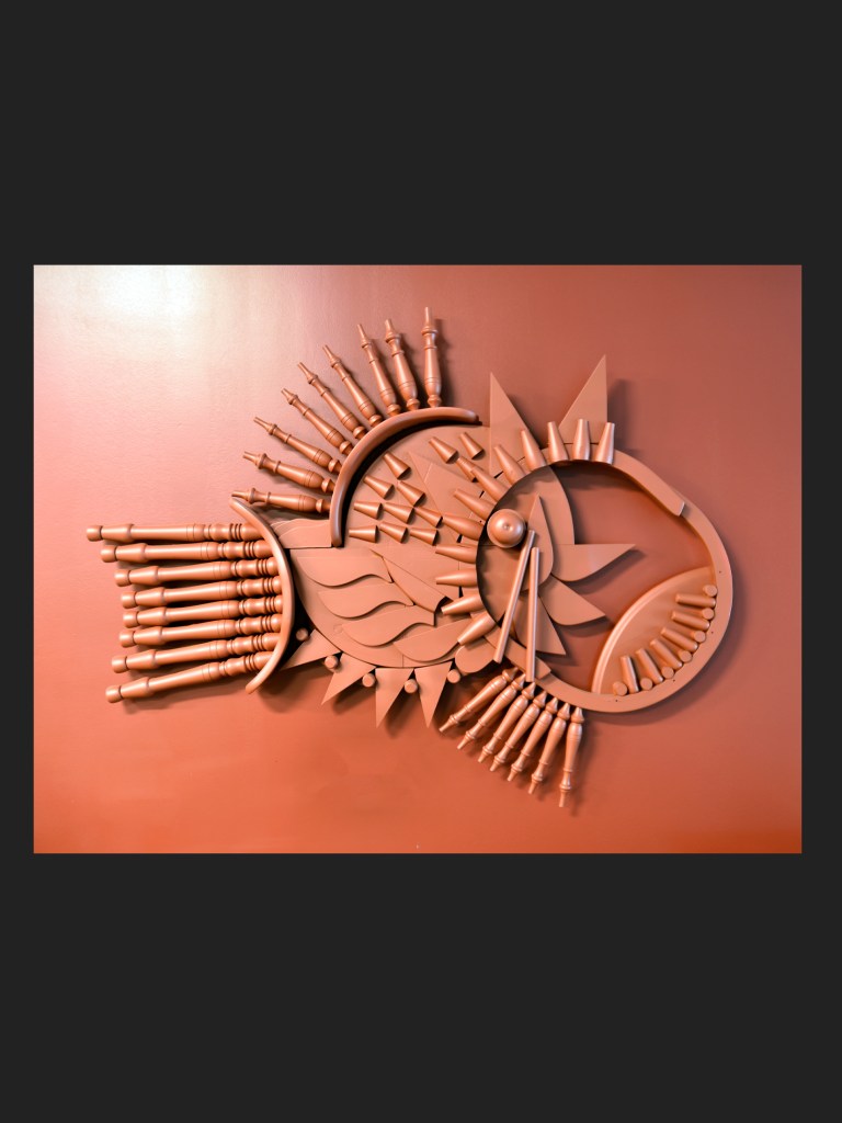 A wall-mounted fish sculpture made from reclaimed wood featuring elongated wooden pieces and geometric shapes, painted in a warm orange color by David Furman