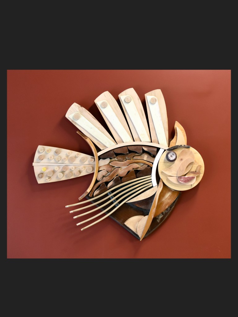 A fish sculpture made from reclaimed wood featuring various shapes and textures, mounted on a red background by David Furman