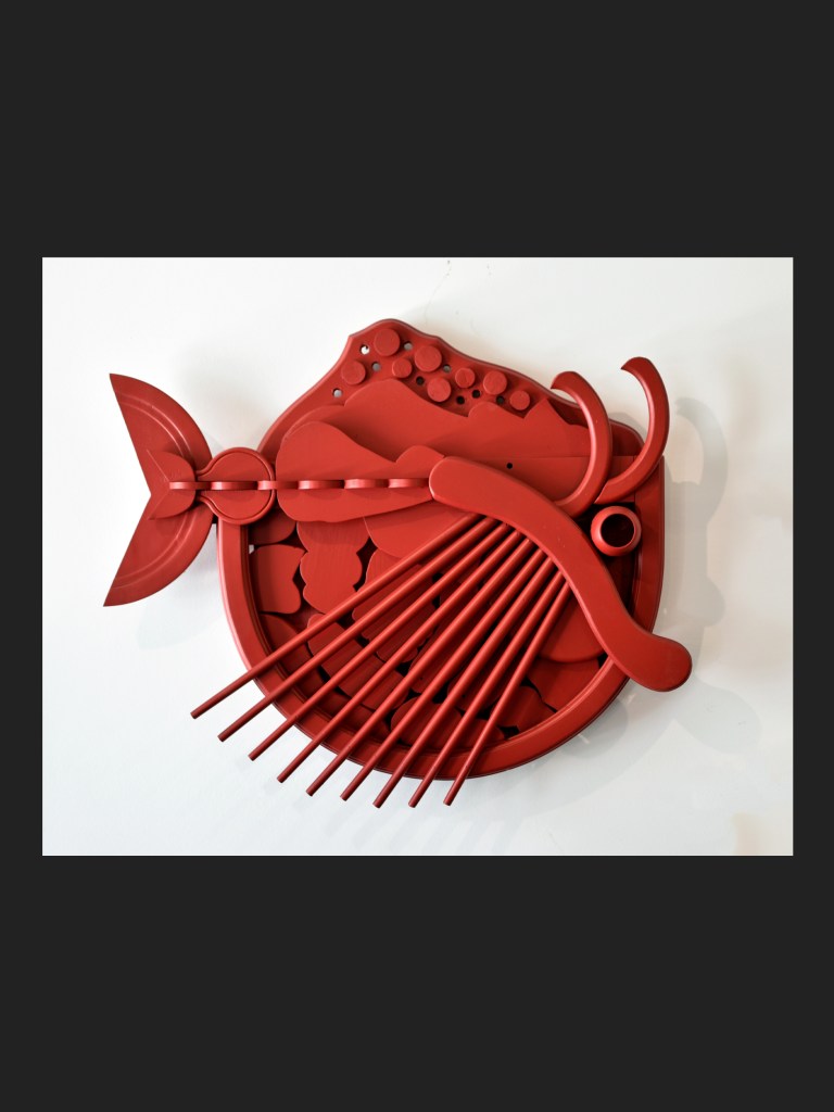 A red artistic representation of a fish made from reclaimed wood with various textured elements and multiple protruding parts, displayed against a neutral background by David Furman