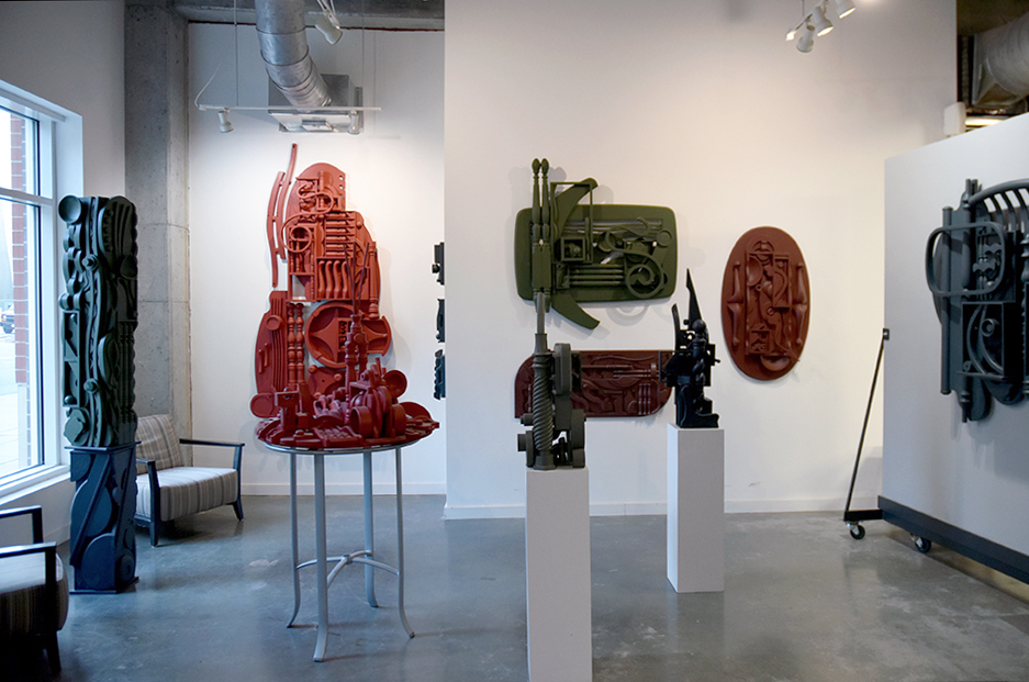 A modern art gallery, David Furman studio, featuring various abstract sculptures in red, green, and black, displayed against white walls and showcasing a variety of artistic forms as pedestals, totems, and wall sculptural hangings, all by David Furman