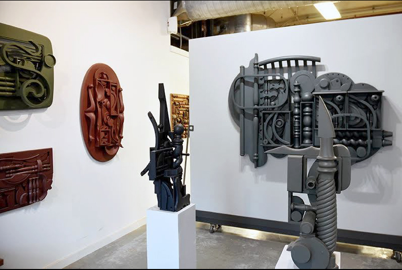 The inside of the David Furman studio and modern art exhibition  space featuring abstract sculptures including totems, pedestal pieces and wall sculptures all in various colors, including red, green, and gray, displayed on white walls, all by David Furman