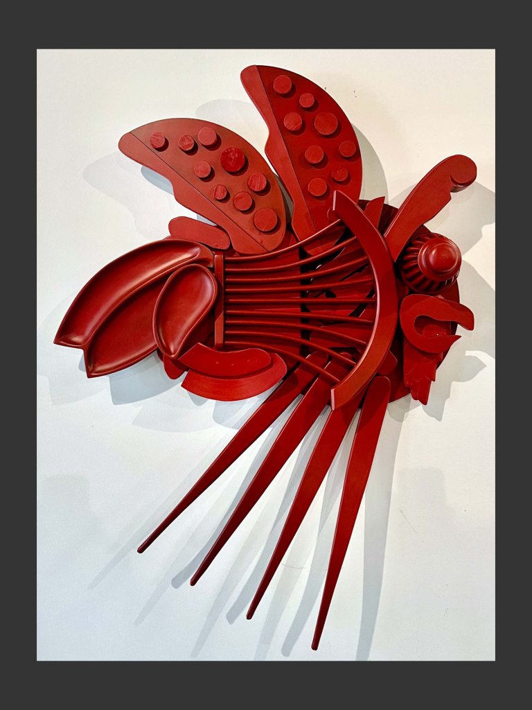 A red wall sculpture resembling a fish made from reclaimed wood with various shapes and textures painted red by David Furman.