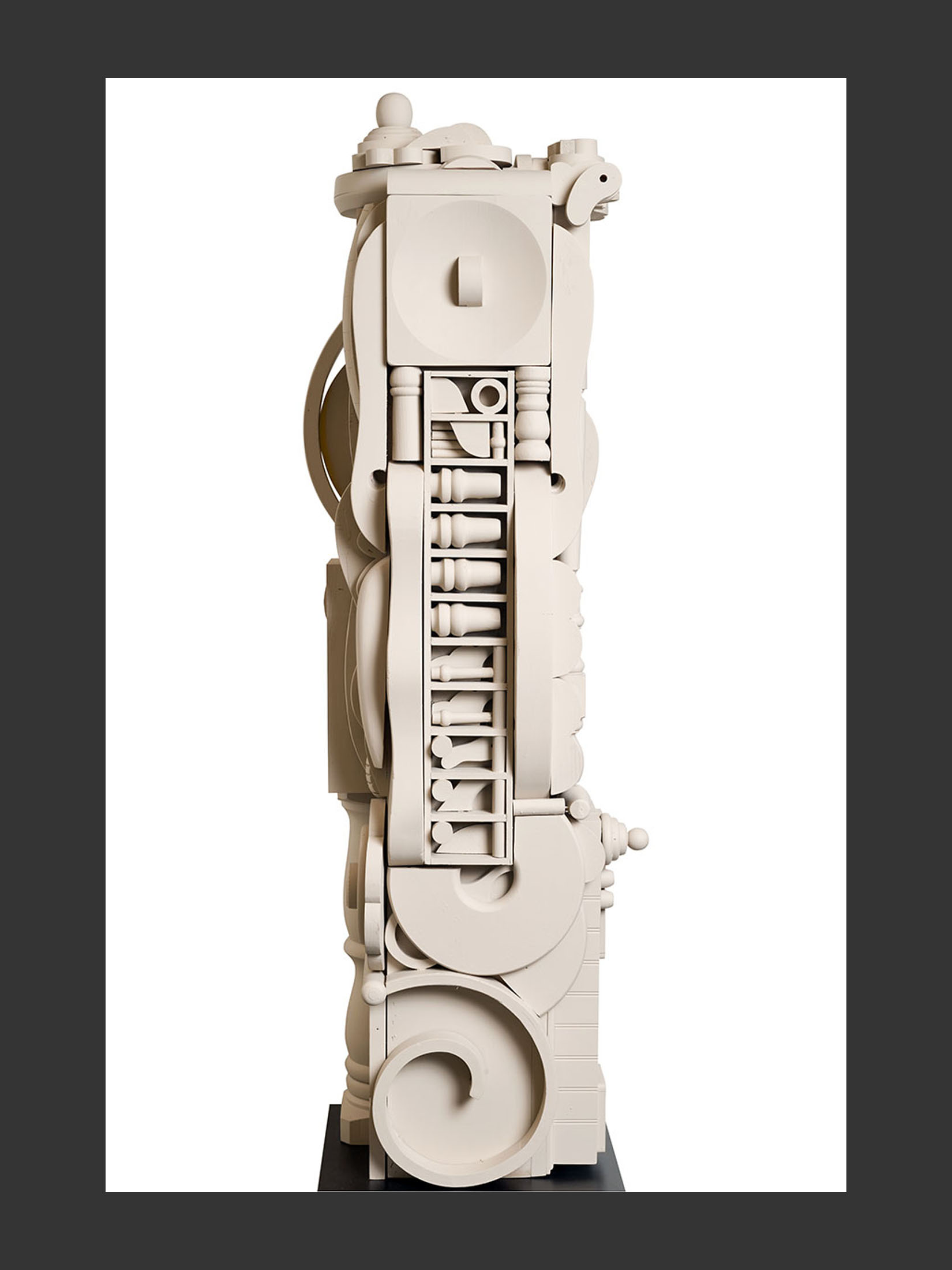 A tall, abstract sculpture (“Totem”) made of repurposed wood featuring a variety of circular and geometric shapes, painted in a light color, and displayed on a black base by David Furman.