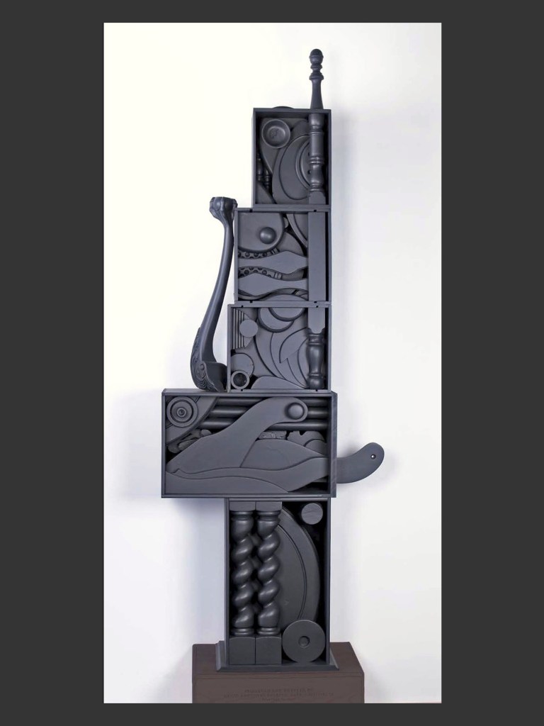 A tall abstract pedestal sculpture made from reclaimed wooden elements composed of darkly colored geometric shapes and forms, featuring cavities and protrusions, presented against a white background by David Furman