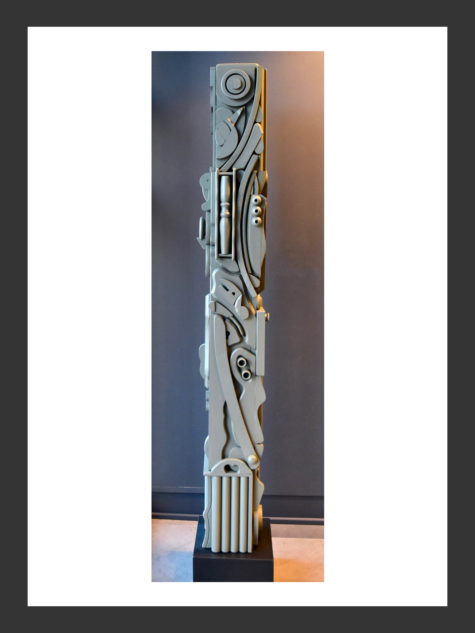 A tall, abstract totem sculpture made from reclaimed wood with intricate patterns and shapes, featuring a gray color scheme, displayed against a dark background on a pedestal by David Furman