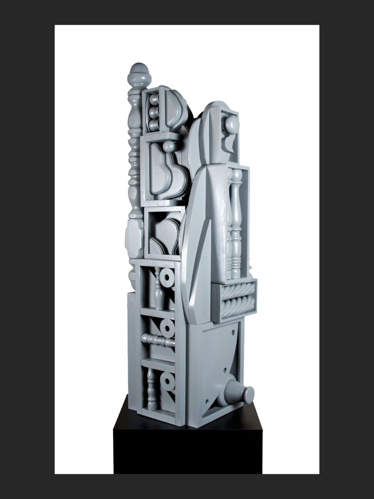 A contemporary sculptural piece in gray made from reclaimed wood, featuring abstract forms and geometric shapes, displayed on a black pedestal by David Furman.