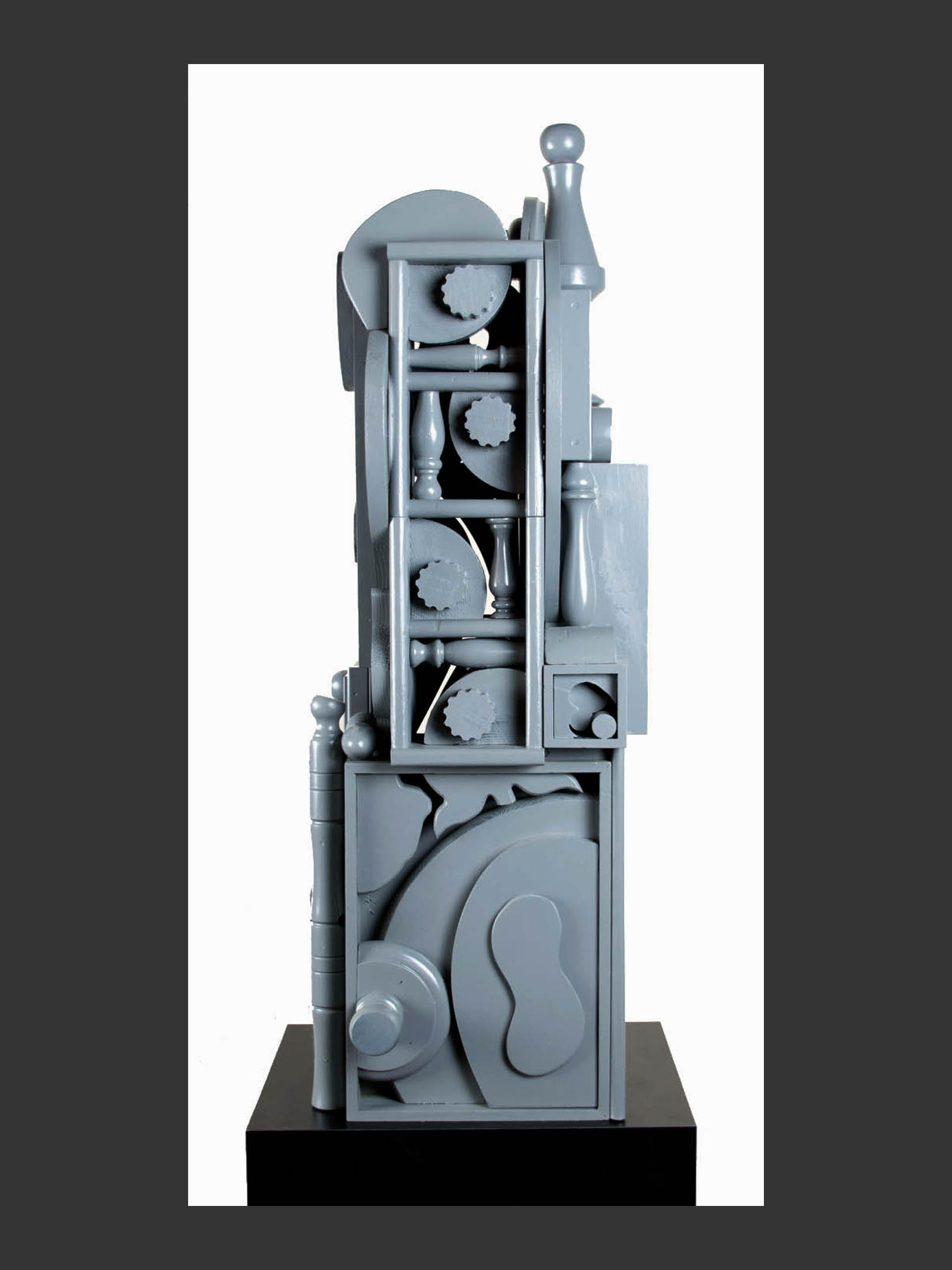 A side view of a large abstract sculpture made from reclaimed wood and composed of various geometric shapes in a uniform gray color, displayed on a dark pedestal against a black background by David Furman.