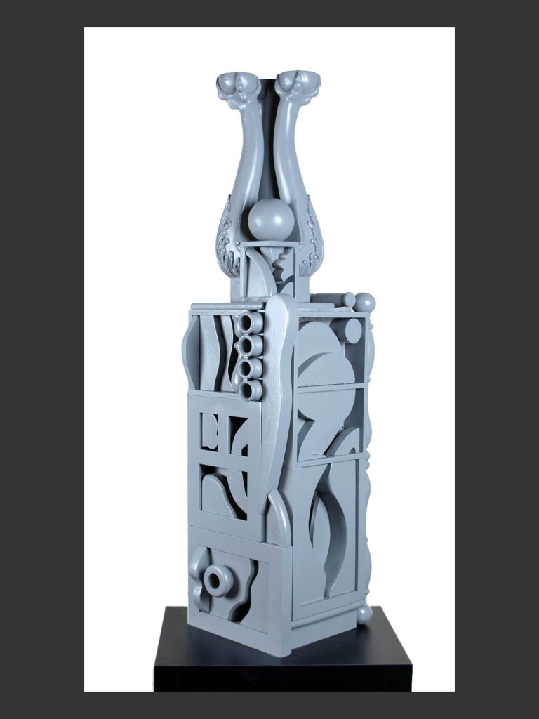 A gray abstract sculpture made from reclaimed wood with two elongated, curved figures at the top, balanced on a blocky base featuring various geometric shapes and cutouts by David Furman.