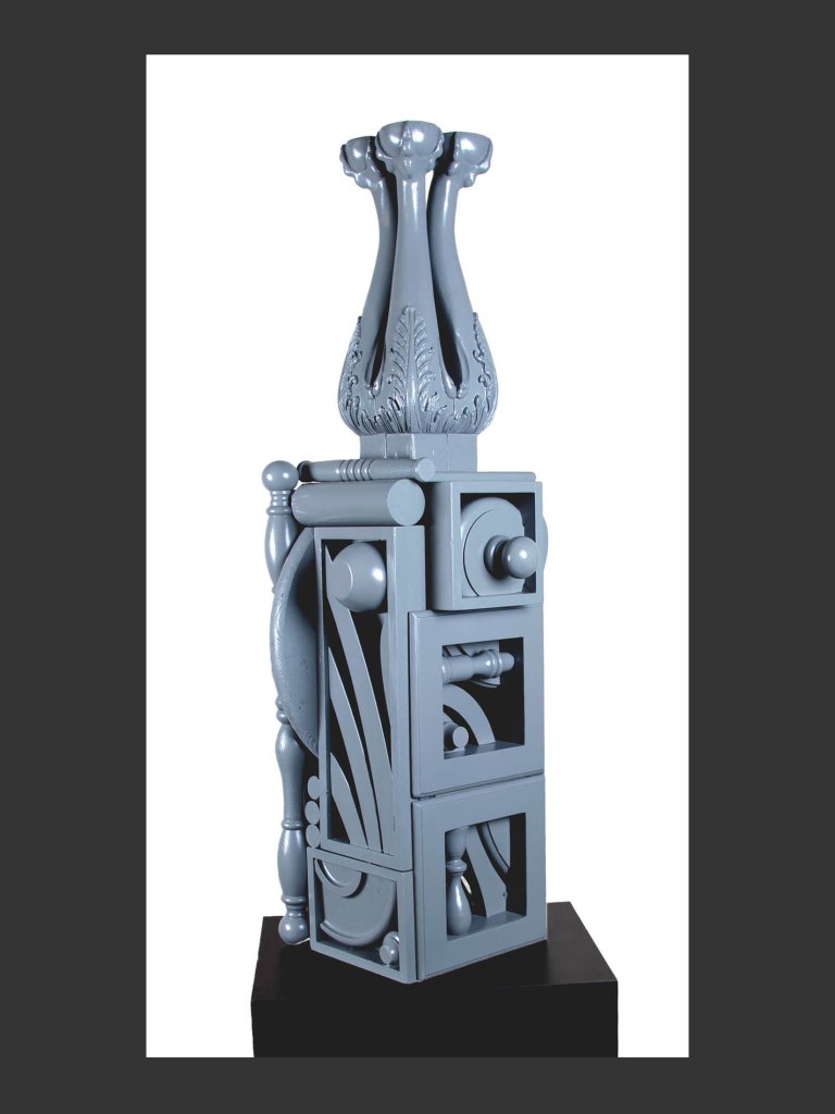 A gray abstract sculpture made from reclaimed wood with two elongated, curved figures at the top, balanced on a blocky base featuring various geometric shapes and cutouts by David Furman.