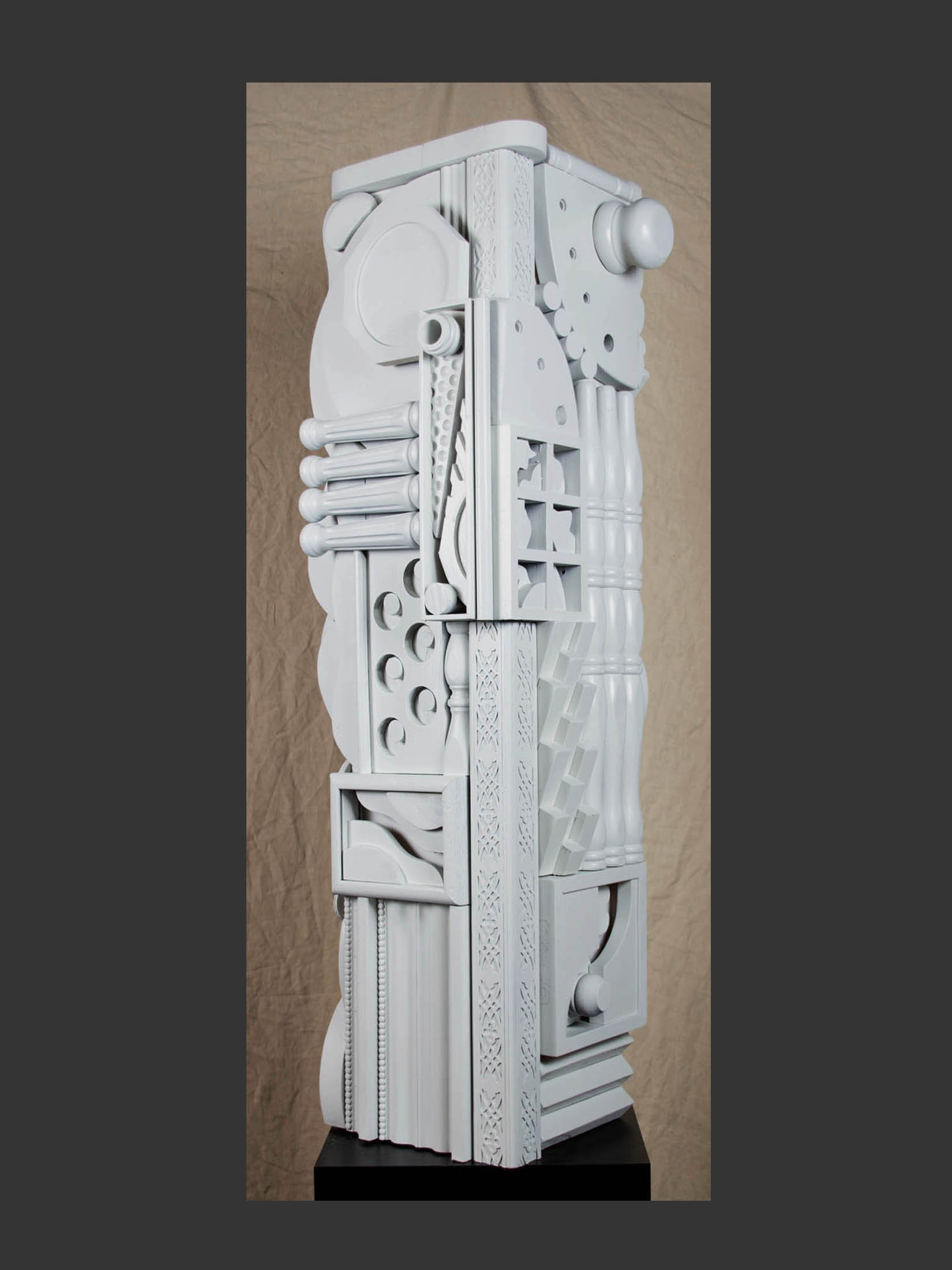 A tall, abstract sculpture made from reclaimed wood and composed of various geometric shapes and patterns, predominantly featuring a white color palette by David Furman.