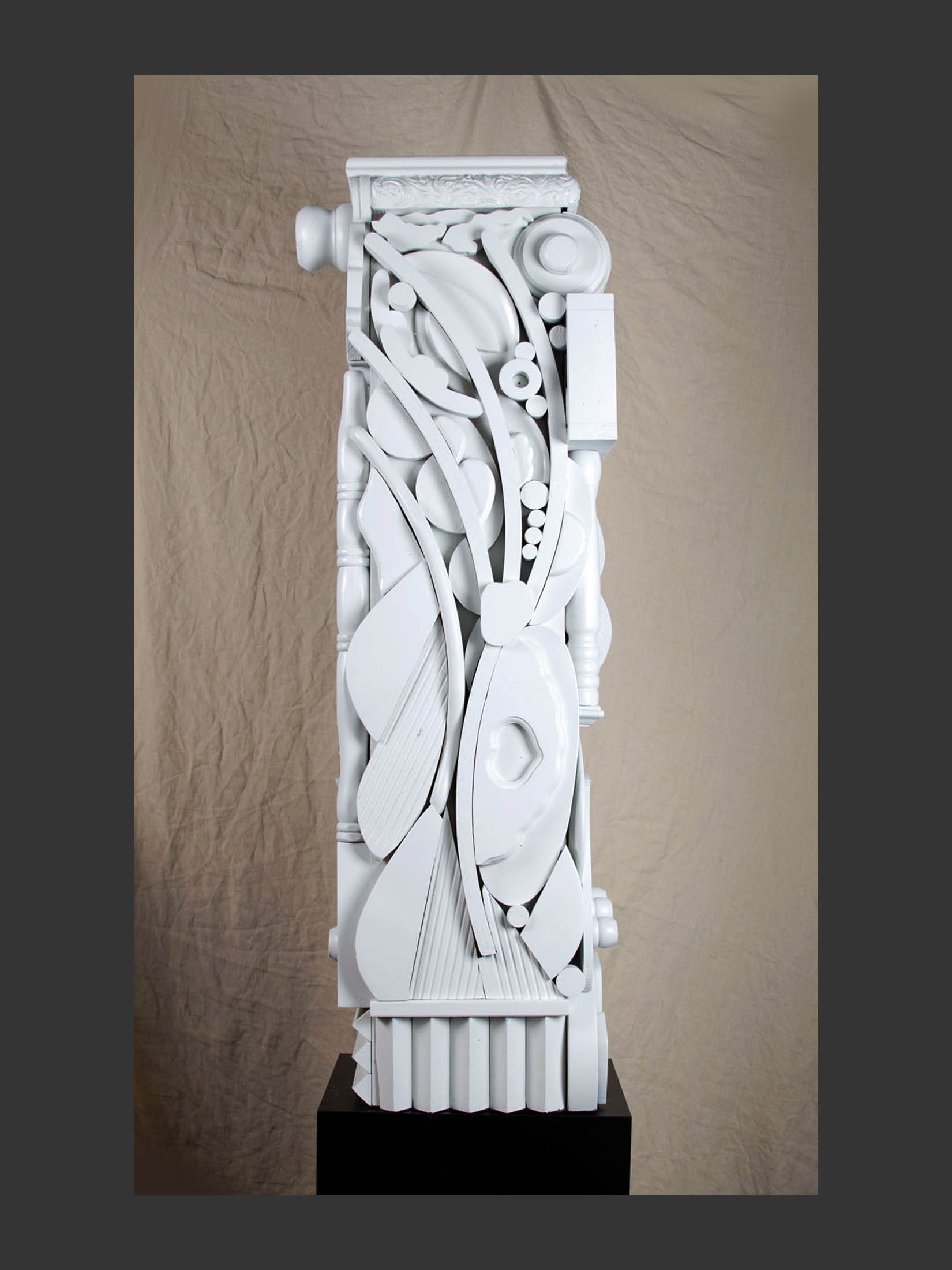 A tall, white abstract sculpture made from reclaimed wood displaying a variety of textured elements arranged in a flowing design, set against a neutral backdrop by David Furman.