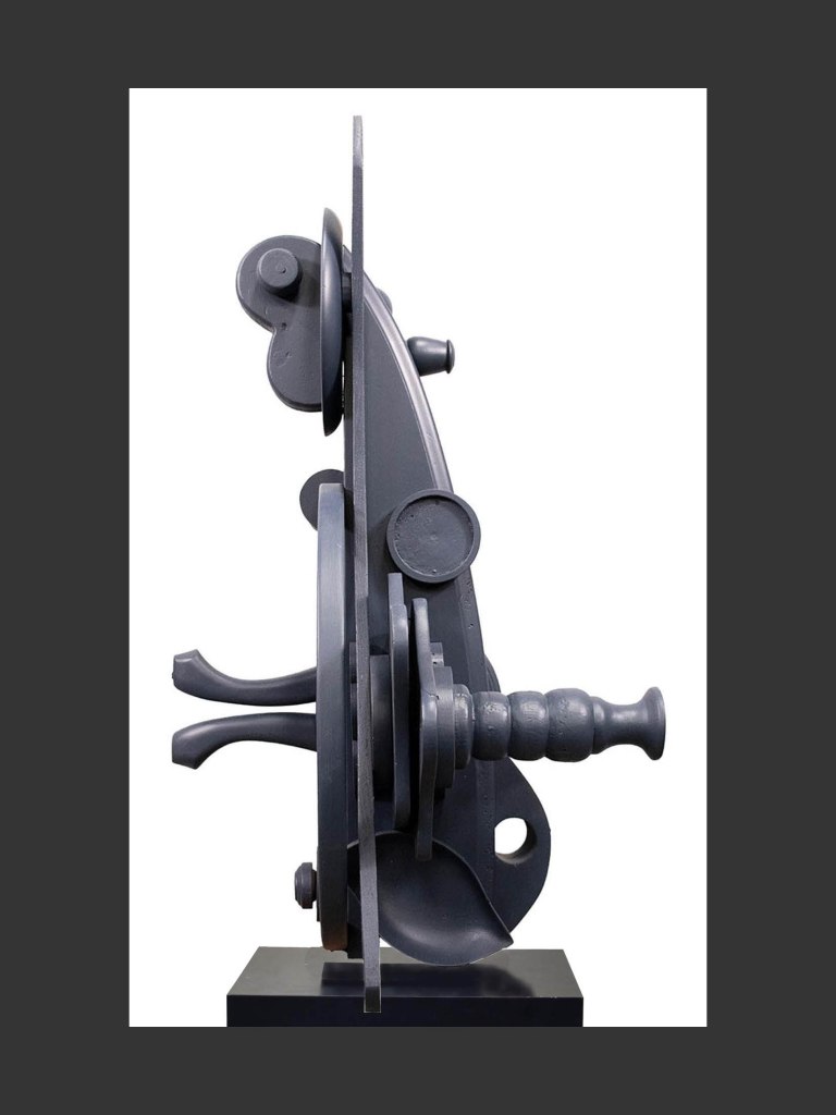 A side profile view of a dark pedestal sculpture made from reclaimed wood featuring geometric components and abstract shapes, displayed on a black pedestal by David Furman