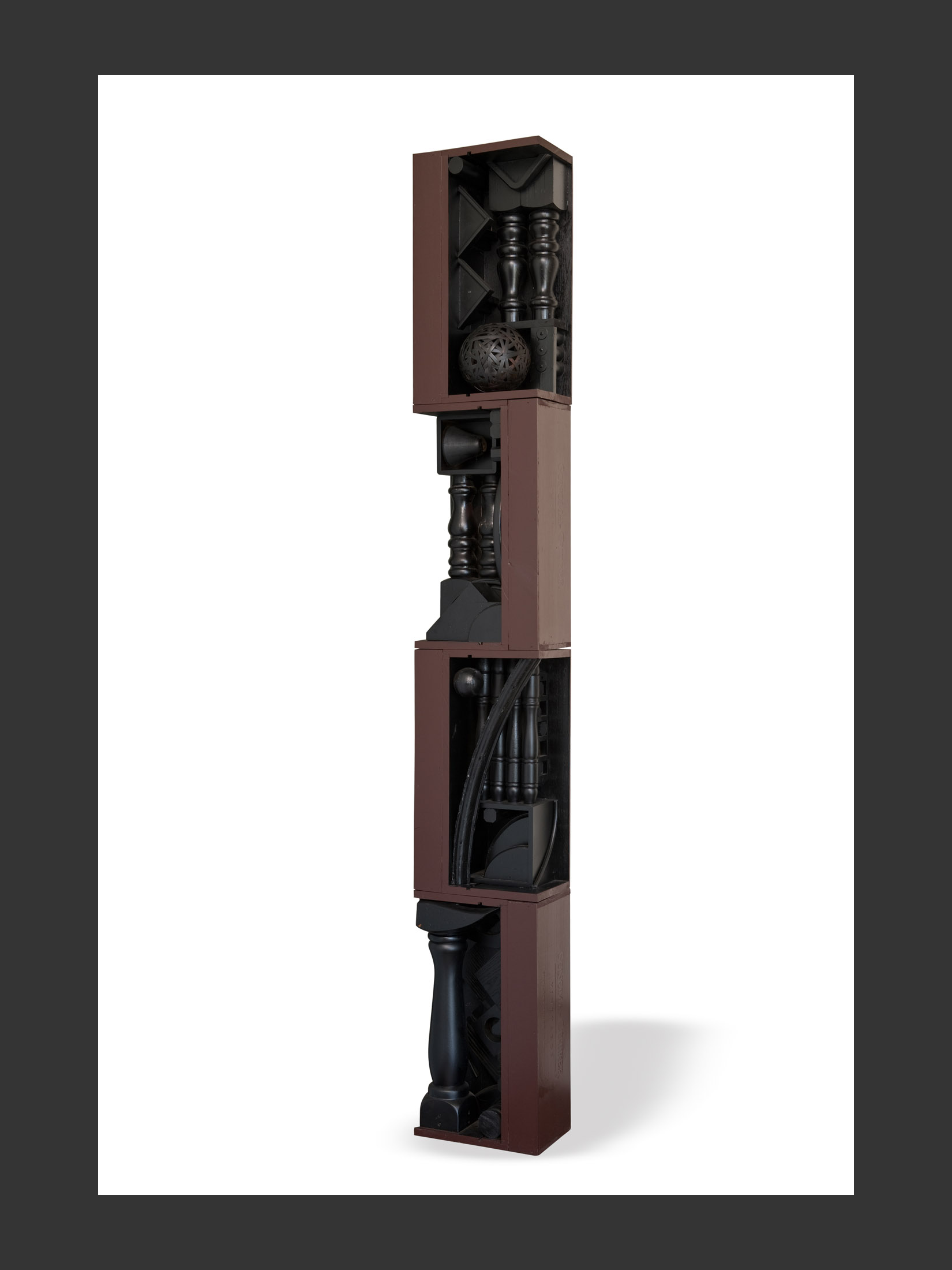 A vertical “Totem” sculpture made from reclaimed wooden elements of stacked rectangular boxes, containing various geometric objects and shapes that resemble architectural elements and painted by David Furman