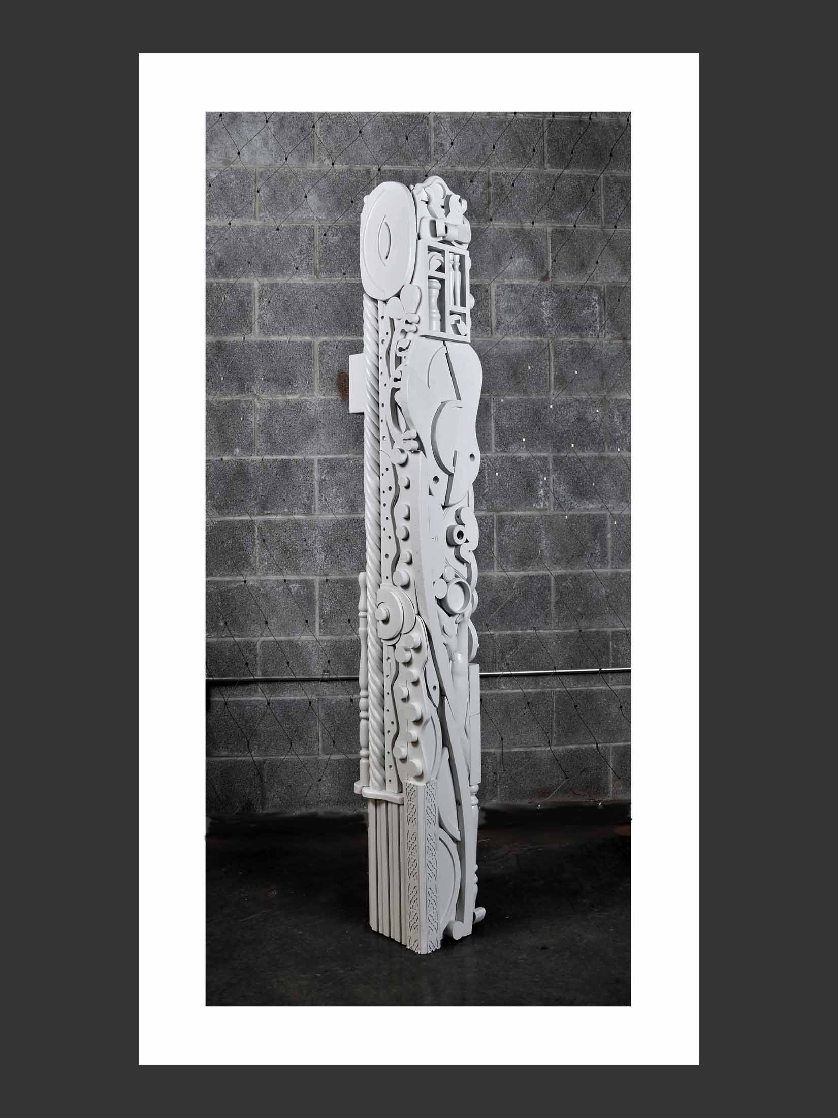 A tall, artistic totem sculpture in white, featuring abstract shapes and intricate designs, set against a textured gray wall by David Furman