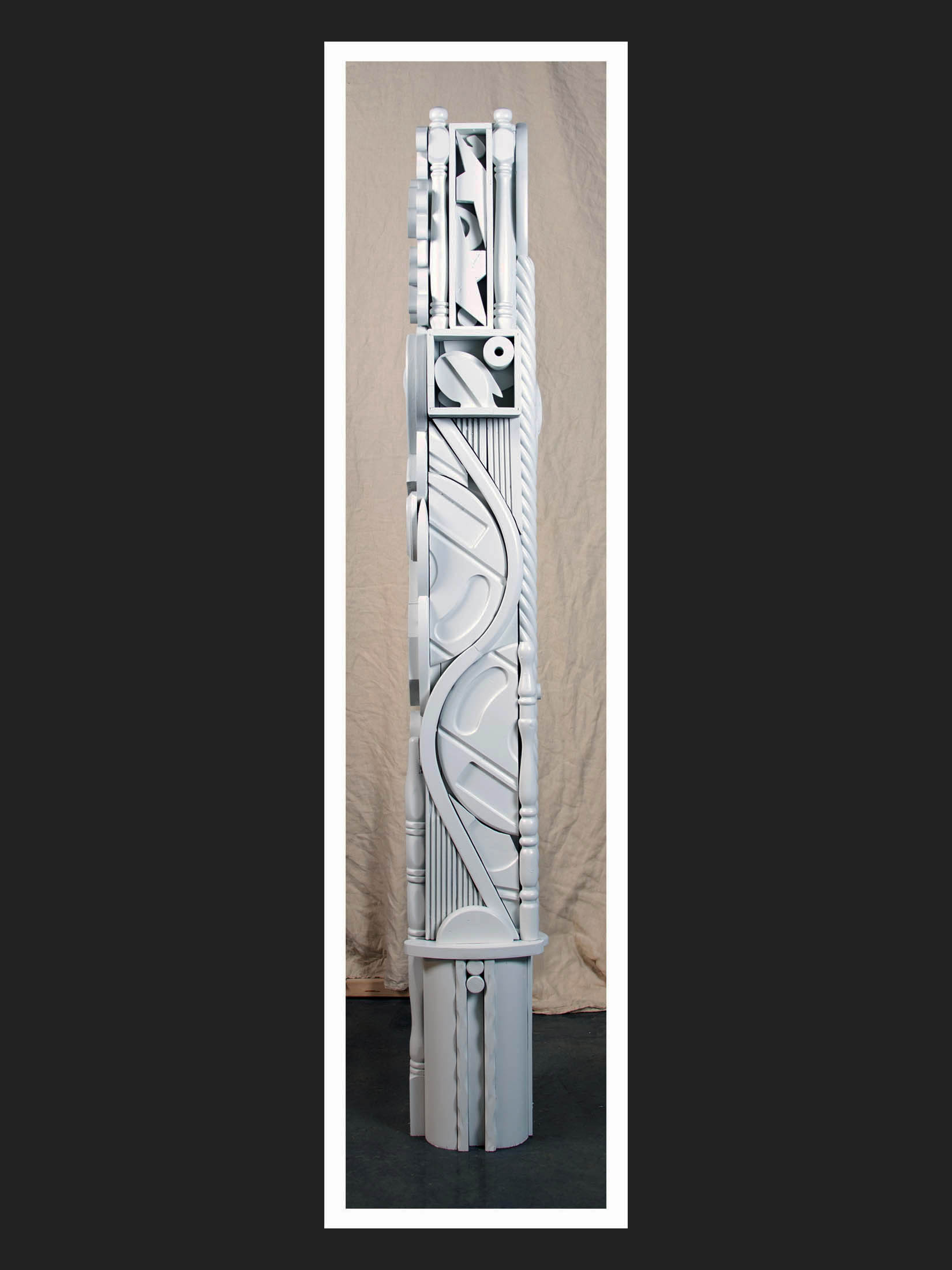 A tall, intricately designed white sculpture Or “Totem”, made of repurposed wooden objects featuring wavy and geometric patterns painted in white by David Furman