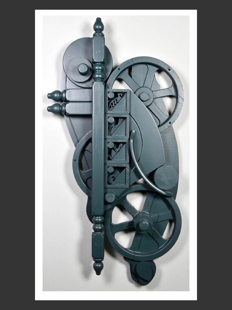 An abstract sculpture made from reclaimed wood featuring a combination of gears, wheels, and other decorative element, all in a gray color scheme by David Furman.